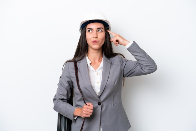 Young architect caucasian woman with helmet and holding blueprints isolated on white background having doubts and thinking