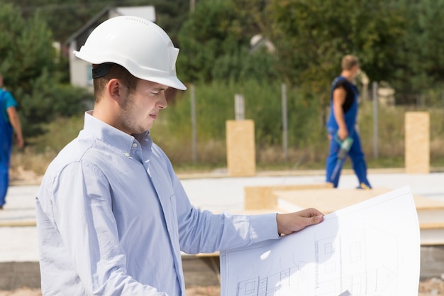 Молодой архитектор или инженер проверяет спецификации на плане или чертеже, стоя с видом на строительную площадку