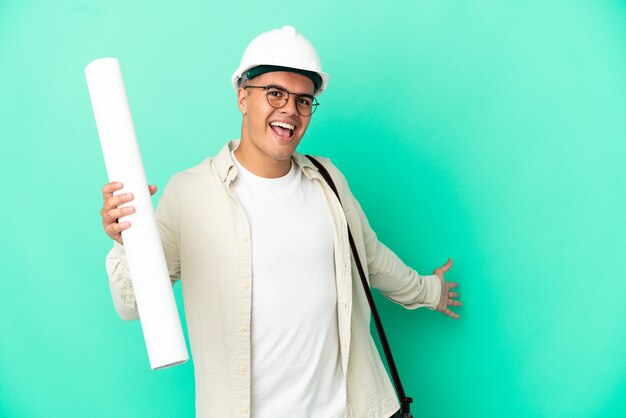 Young architect man holding blueprints over isolated background extending hands to the side for inviting to come