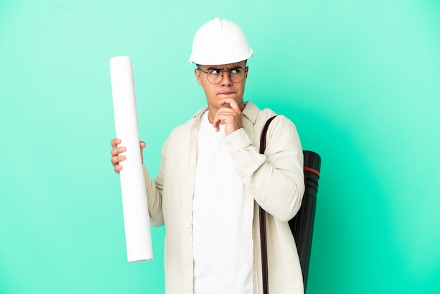 Young architect man holding blueprints over isolated background having doubts and thinking