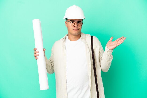 Young architect man holding blueprints over isolated background having doubts while raising hands