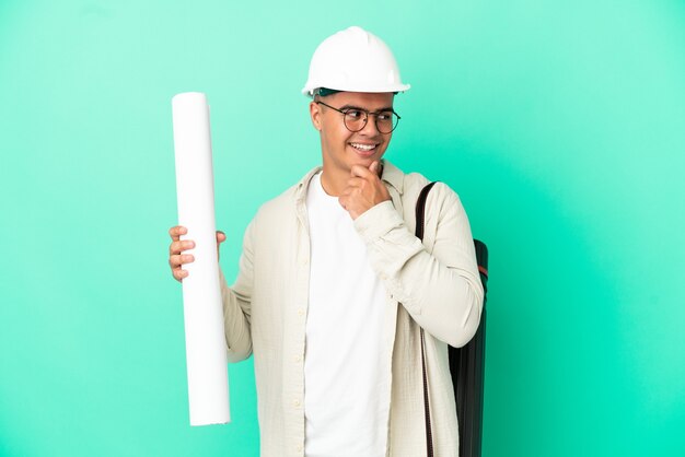 Young architect man holding blueprints over isolated background looking to the side and smiling