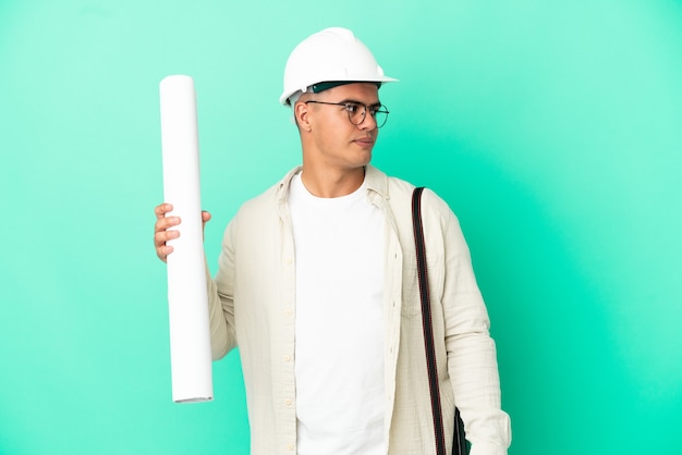 Young architect man holding blueprints over isolated background looking to the side