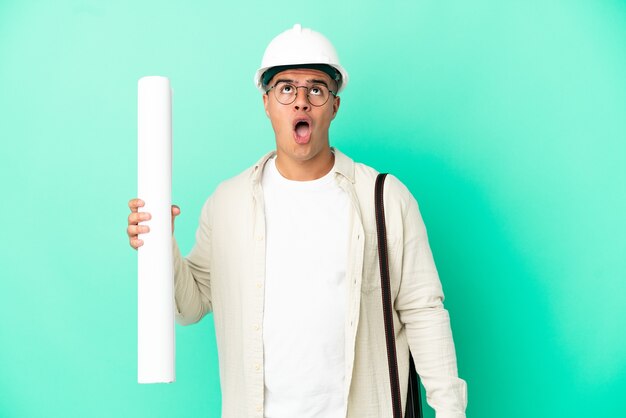 Young architect man holding blueprints over isolated background looking up and with surprised expression