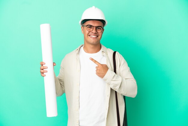 Young architect man holding blueprints over isolated background pointing to the side to present a product