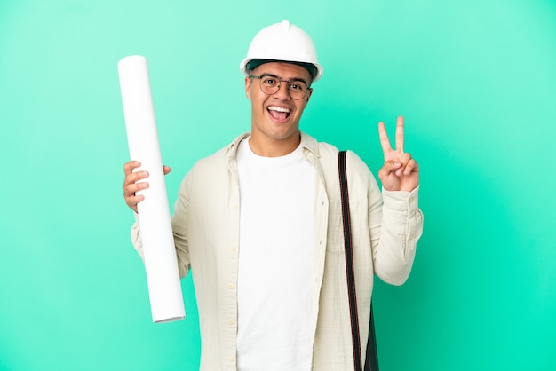 Young architect man holding blueprints over isolated background smiling and showing victory sign