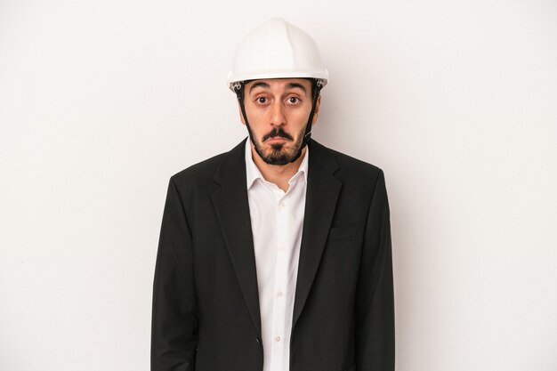 Young architect man wearing a construction helmet isolated on white background shrugs shoulders and open eyes confused.