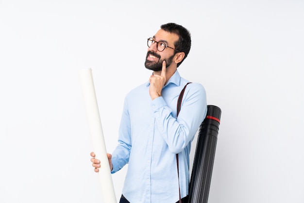 Giovane uomo dell'architetto con la barba sopra la parete bianca isolata che pensa un'idea mentre osservando in su