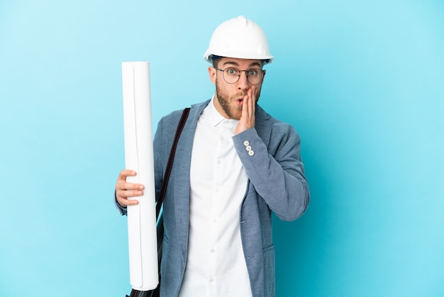 Young architect man with helmet and holding blueprints over isolated background with surprise and shocked facial expression