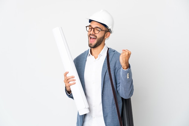 Young architect man with helmet and holding blueprints isolated on white wall celebrating a victory