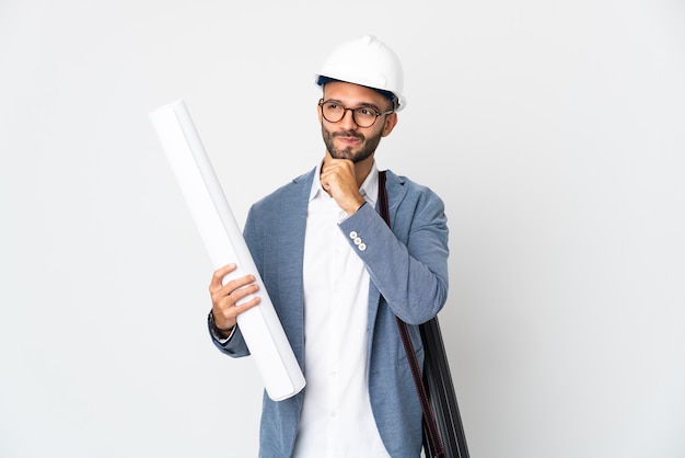 Young architect man with helmet and holding blueprints isolated on white wall having doubts and thinking