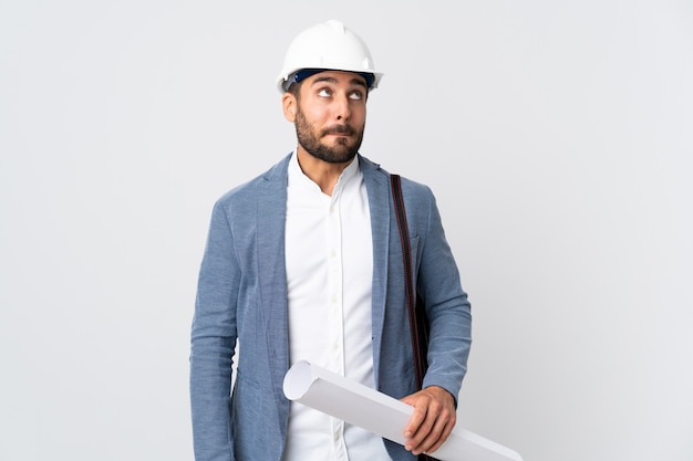 Young architect man with helmet and holding blueprints isolated on white wall having doubts while looking up