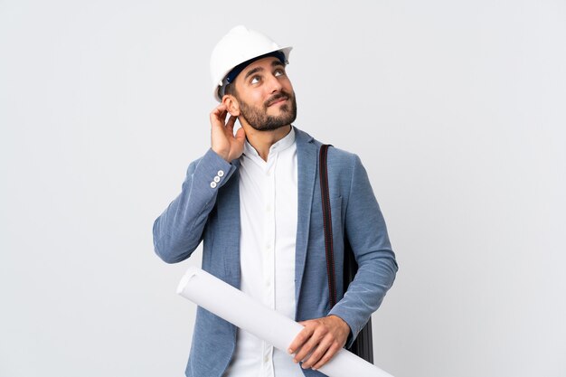 Young architect man with helmet and holding blueprints isolated on white wall thinking an idea