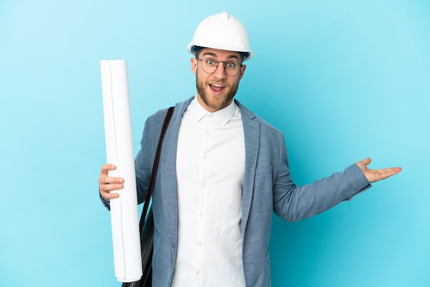 Young architect man with helmet and holding blueprints over isolated with shocked facial expression