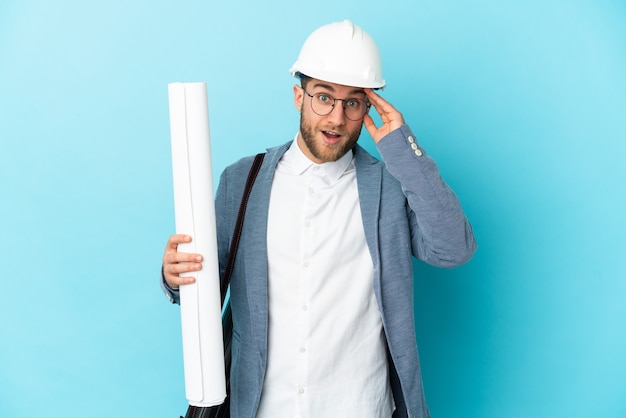 Young architect man with helmet and holding blueprints with surprise expression