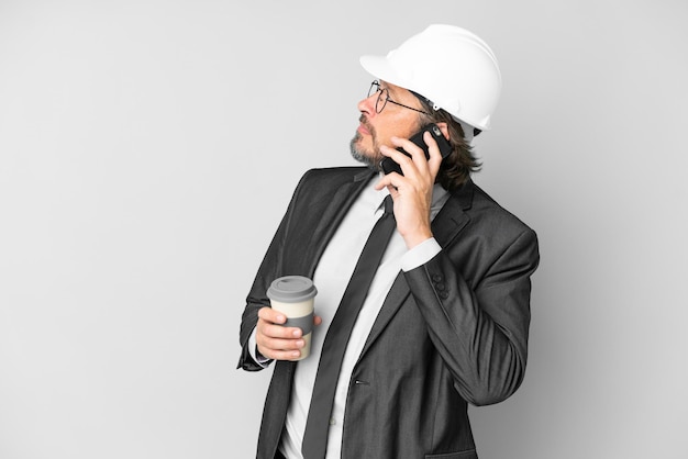 Young architect man with helmet over isolated background holding coffee to take away and a mobile