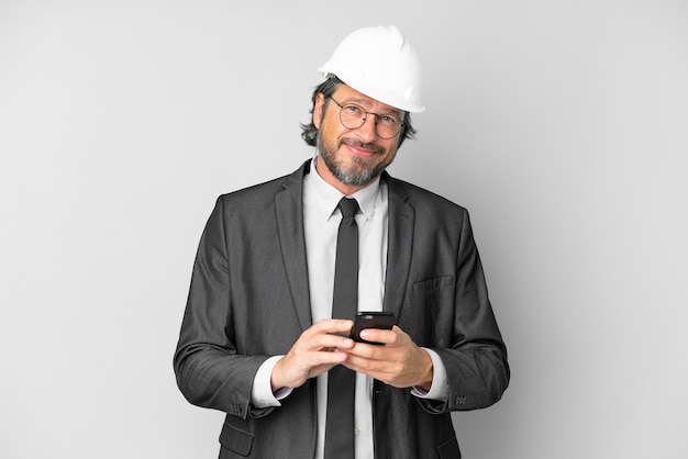 Young architect man with helmet over isolated background sending a message with the mobile