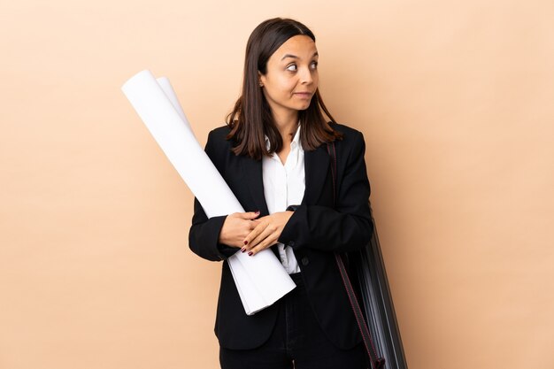 Young architect woman holding blueprints over isolated making doubts gesture looking side