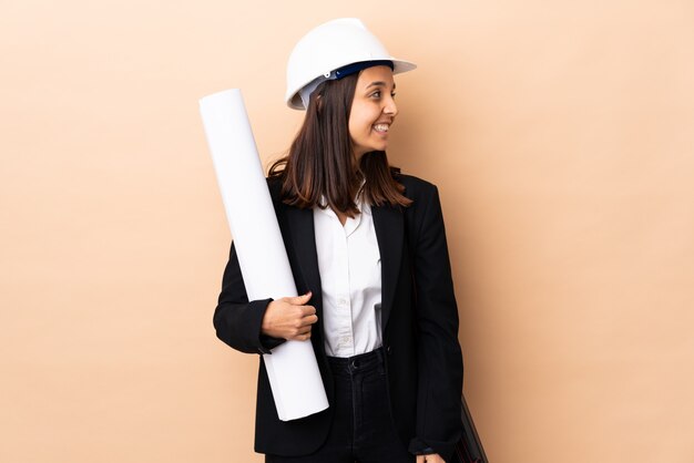 Young architect woman holding blueprints over isolated wall looking side