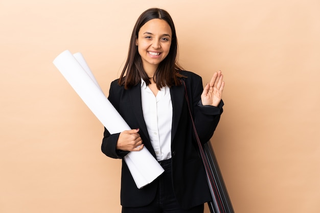 Young architect woman holding blueprints over wall saluting with hand with happy expression