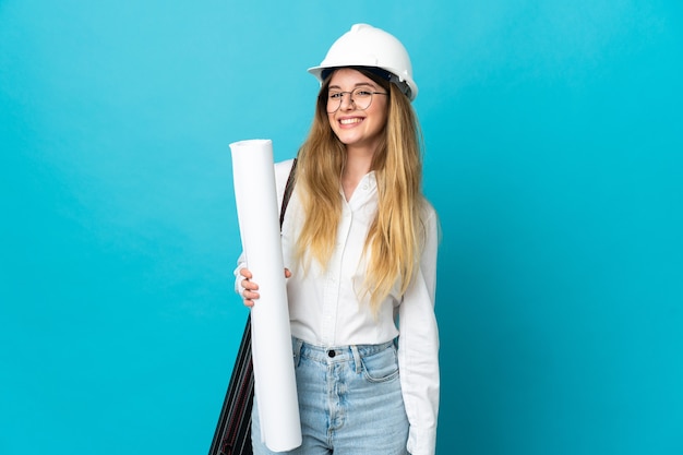 Young architect woman with helmet and holding blueprints on blue looking to the side and smiling