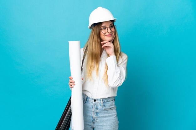 Young architect woman with helmet and holding blueprints isolated on blue wall thinking an idea while looking up