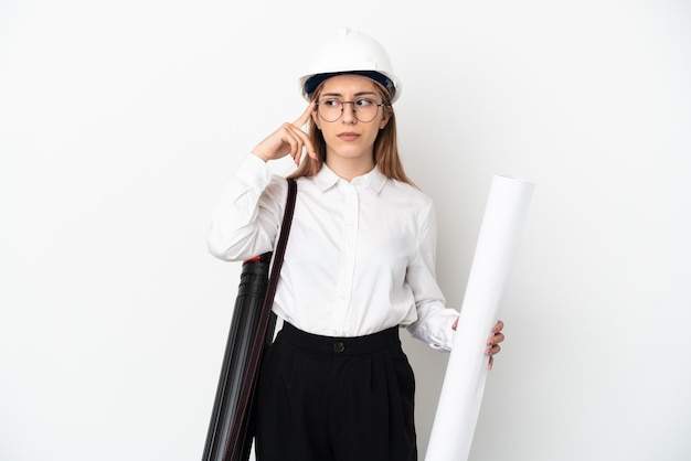 Young architect woman with helmet and holding blueprints isolated on white background having doubts and thinking