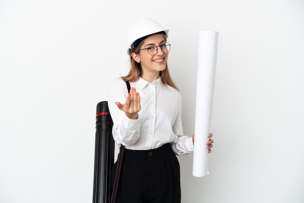 Young architect woman with helmet and holding blueprints isolated on white wall inviting to come with hand. Happy that you came