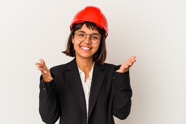 Young architect woman with red helmet isolated on white background feels confident giving a hug to the camera.