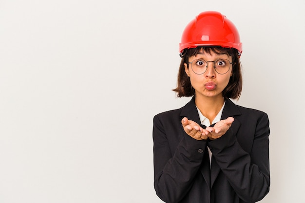 Young architect woman with red helmet isolated on white background folding lips and holding palms to send air kiss.