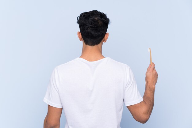 Young Argentinian man brushing his teeth over isolated blue wall in back position