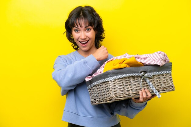 Young argentinian man holding a clothes basket isolated on yellow background celebrating a victory