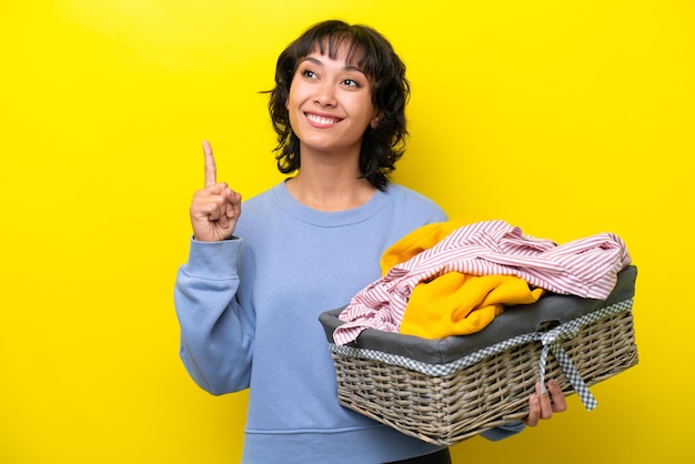 素晴らしいアイデアを指している黄色の背景に分離された服のバスケットを保持している若いアルゼンチン人