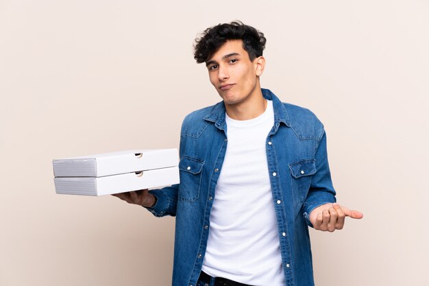 Young Argentinian man holding pizzas over isolated wall making doubts gesture while lifting the shoulders