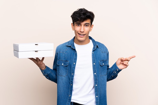 Young Argentinian man holding pizzas over isolated wall surprised and pointing finger to the side