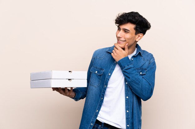 Young Argentinian man holding pizzas over isolated wall thinking an idea and looking side