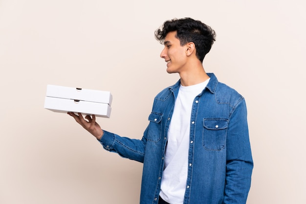 Молодой аргентинский мужчина держит пиццу на изолированной стене с счастливым выражением