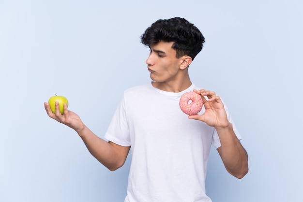 Young Argentinian man over isolated blue wall having doubts while taking a chocolate tablet in one hand and an apple in the other