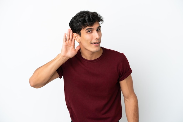 Young Argentinian man isolated on white background listening to something by putting hand on the ear