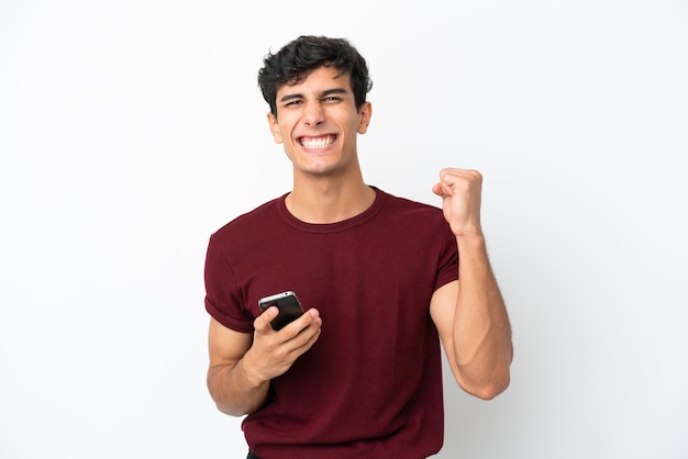 Young Argentinian man isolated on white background with phone in victory position