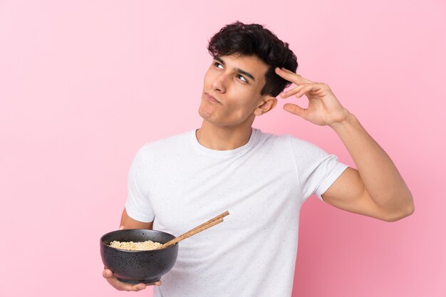 Young Argentinian man over isolated white wall having doubts and with confuse face expression while holding a bowl of noodles with chopsticks