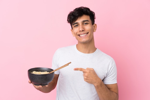 Young Argentinian man over isolated white wall and pointing it while holding a bowl of noodles with chopsticks