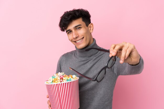 Young Argentinian man over isolated white wall with 3d glasses and holding a big bucket of popcorns