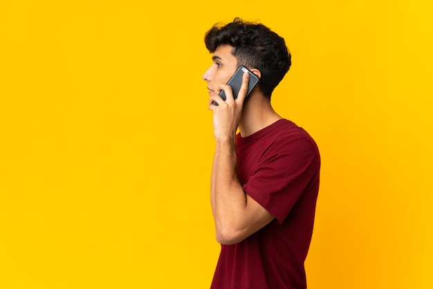 Young Argentinian man isolated on yellow background keeping a conversation with the mobile phone with someone