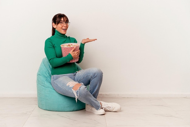 Young Argentinian woman eating popcorns on a puff isolated on white background showing a copy space on a palm and holding another hand on waist.