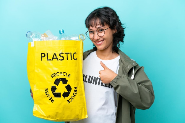 Young argentinian woman holding a bag full of plastic bottles to recycle isolated on blue background giving a thumbs up gesture