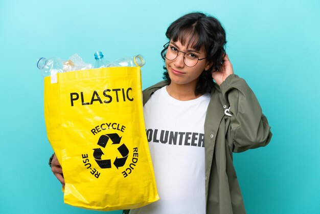 Young Argentinian woman holding a bag full of plastic bottles to recycle isolated on blue background having doubts