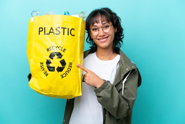 Young Argentinian woman holding a bag full of plastic bottles to recycle isolated on blue background pointing to the side to present a product