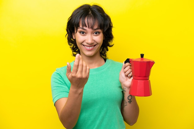 Young argentinian woman holding coffee pot isolated on yellow background inviting to come with hand happy that you came