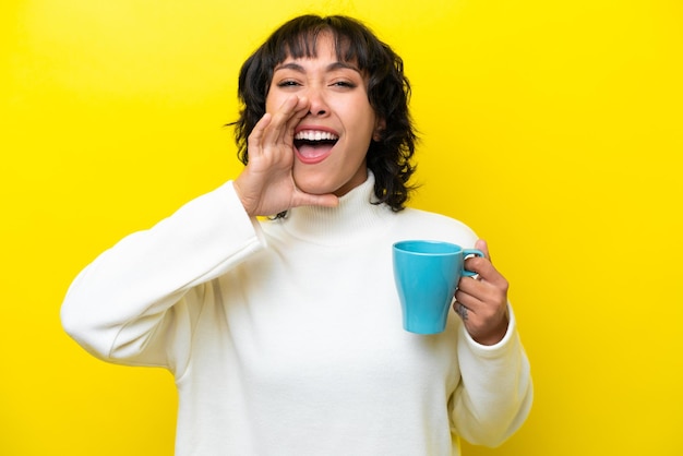 Young Argentinian woman holding cup of coffee isolated on yellow background shouting with mouth wide open